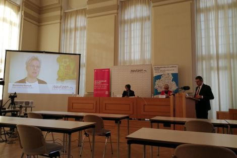 Pressekonferenz, 2 Männer und 1 Frau. Ein Mann redet am Podest. Im Hintergrund wird eine Video gezeigt.