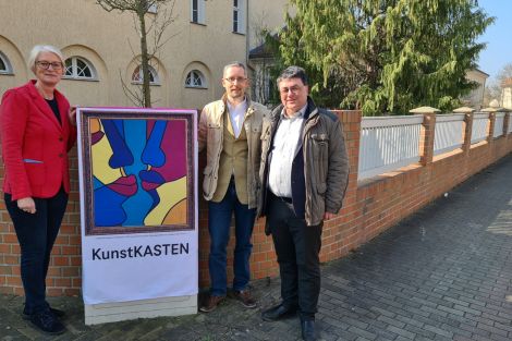 3 Personen zusammen vor einem zum Kunstkasten verhüllten Netzverteiler vor einer Mauer in Lauta.